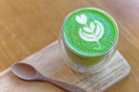Matcha latte with heart-shaped latte art in clear glass on wooden tray. Hot matcha latte with delicate heart and leaf latte art served in double-wall glass cup on wooden tray with a spoon on the sideの写真素材