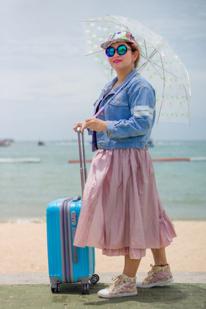 Stylish Woman with Luggage and Umbrella at a Tropical Beach. A fashionable woman in a denim jacket, pink skirt, and sunglasses stands with a bright blue suitcase and a clear umbrella on a beach.の写真素材