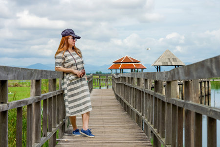 Woman Enjoying Scenic View at Bueng Bua Wood Boardwalk. Tourist stands on rustic wooden boardwalk at Bueng Bua, Sam Roi Yot, taking in serene wetland landscape under cloudy skyの写真素材