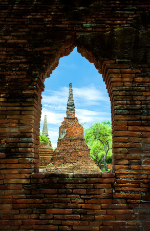 View of Historic Thai Pagodas Through Ancient Brick Arched Window. Unique perspective of ancient brick chedis at Wat Phra Si Sanphet, framed by a weathered red brick window.の写真素材