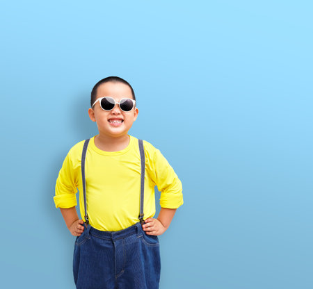 A little boy is hip-akimbo posting and wearing big sunglasses. Isolated on a blue background,の写真素材