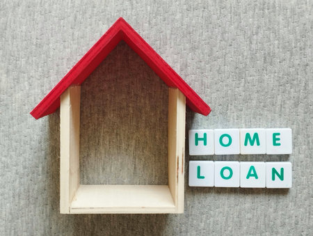 A simple wooden house model with a red roof placed next to the words "HOME LOAN" in letter tiles on a gray textured background, representing real estate financing and mortgages.の写真素材