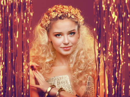Beautiful young girl with blond curly hair, wearing golden dress and golden accessories standing behind a curtain made of golden ribbonsの写真素材