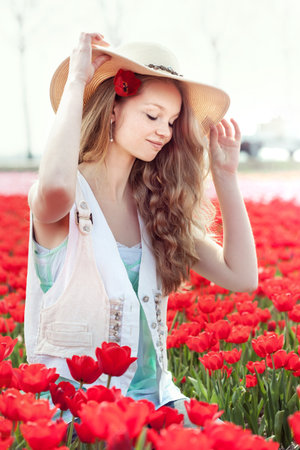 beautiful young woman with hat in her hands on the meadow with red flowers tulips, outdoorsの写真素材
