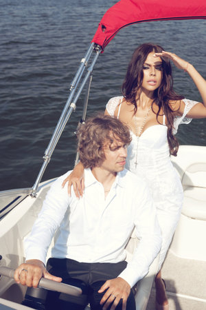 Beauty lovely couple posing on the luxury boat in open sea in summer. Young man and sensual brunette outdoor portrait in classic dress. Outdoors, lifestyle.の写真素材