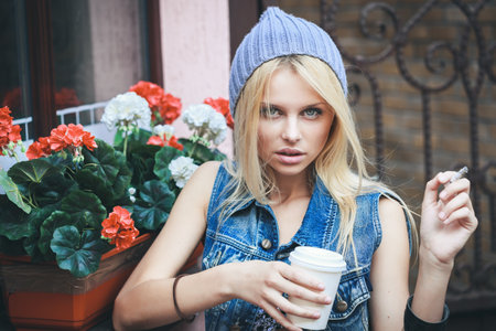 Young pretty sensual blonde woman sitting outdoor on the street while smoking sigarette with cup of hot coffee espresso. Outdoors, lifestyle.の写真素材