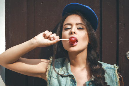 Young sexy girl in blue cap and jeans jacket sucking lollipop.  Outdoors, lifestyle.の写真素材