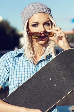 A young woman in sunglasses is standing on the street and holding a skateboard in front of her.  Outdoor lifestyle.の写真素材
