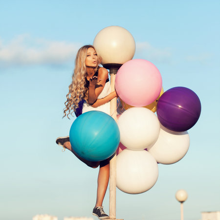 Happy young girl with big colorful latex balloons. Beauty Romantic Girl Outdoors. Woman send air kiss on lamppost on background of blue sky.の写真素材
