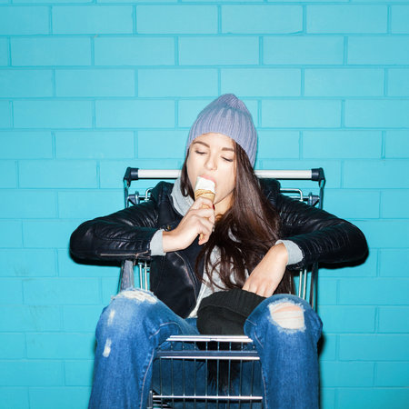 Young funky woman eating ice cream over blue brick wall. Naughty girl having fun in shopping cart....