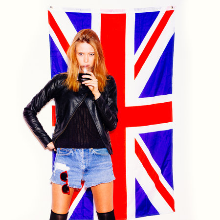 Young woman with glass of alcohol standing overthe Union Jack flag on whiteの写真素材