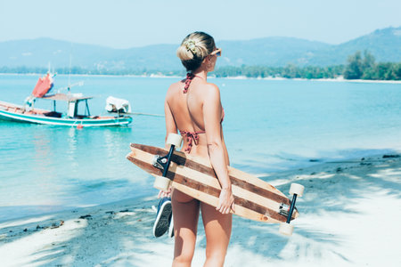 Fashion lifestyle outdoors. Sporty woman with longboard in hand walking on white sand on tropical...