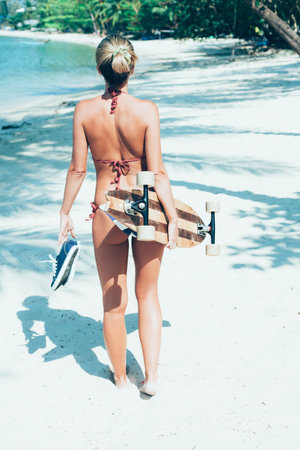 Fashion lifestyle outdoors. Sexy woman with longboard in hand walking on white sand on tropical...