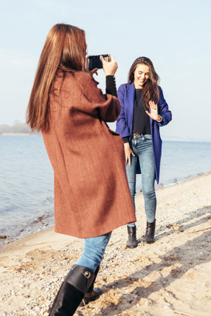 Beautiful young girl taking pictures her cheerful girlfriend on the beach in front of the sea. Outdoor lifestyle portraitの写真素材