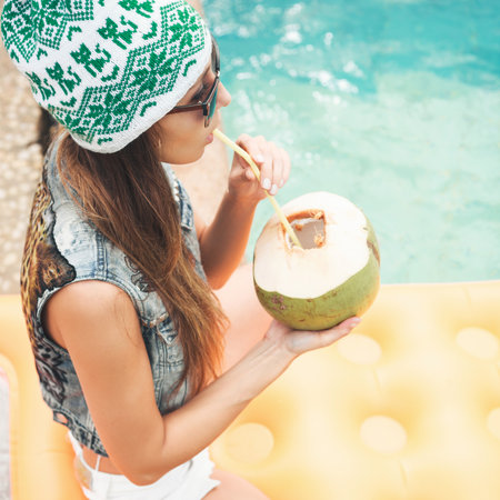 Beautiful young woman in bikini drinking coconut juice. Female legs in the pool water. Outdoor lifestyle portraitの写真素材