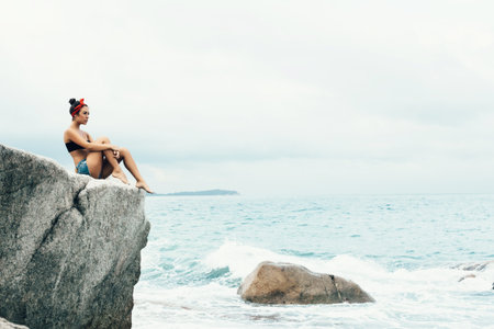 Rear view of an attractive woman sitting and relaxing on a natural coastal rock high up,...
