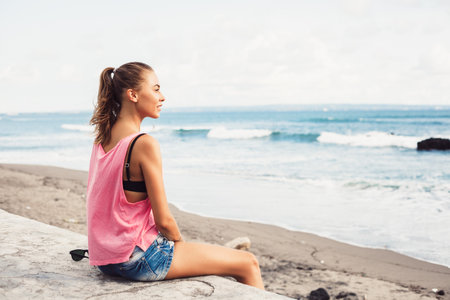 Outdoor fashion portrait summer beach style of Young happy woman sitting near the sea and looking on the water and waiting for somebody in oceanの写真素材