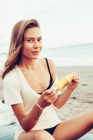 Outdoor summer style fashion portrait of young beautiful sensual woman posing on the beach on vacation. Girl holding corn and smiling. Snacking on the sea.の写真素材