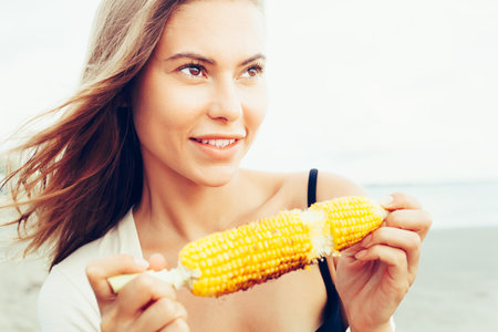 Outdoor summer style fashion portrait of young beautiful sensual woman posing on the beach on vacation. Happy pretty Girl holding corn. Snacking on the sea.の写真素材