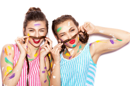 Two lovely girl friends having fun. Women smeared in paint show a mustache from the hair. White background not isolatedの写真素材