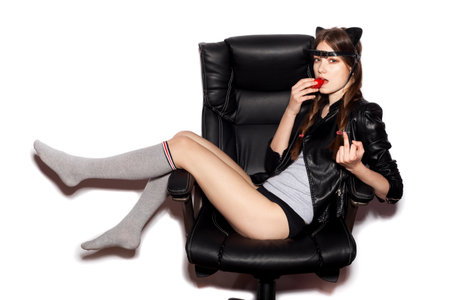 Young woman in cat ears sit on black office chair eat strawberry and show middle finger . Fashion hipster girl.  White background, not isolatedの写真素材