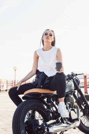 Young sexy girl sitting on vintage custom motorcycle and drinking coffee . Outdoor lifestyle portraitの写真素材