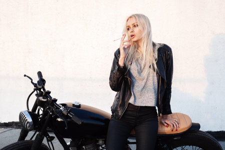 Fashion female biker girl. Young blonde woman sitting on vintage motorcycle and smoking cigarette. Outdoors lifestyle portraitの写真素材