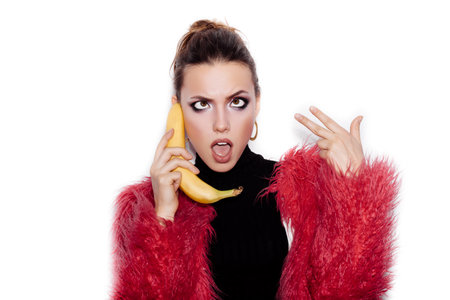 Fashion swag female model wearing black dress and pink fur coat making fun with banana. Woman holding a banana as a telephone over a white background not isolatedの写真素材