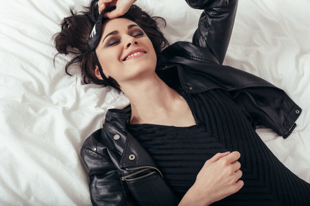The girl with a mask for sleep lying in bed and smiling. Young woman resting in a leather jacketの写真素材