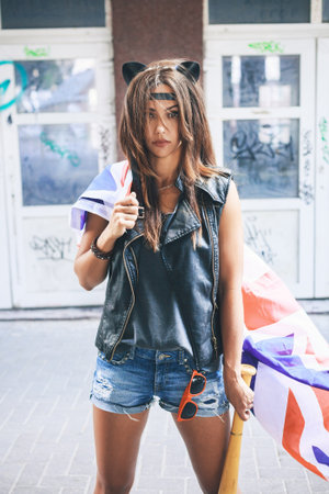 Bully girl with leather cat ears holding a baseball bat and British flag at street. Urban scene. Outdoor lifestyle portraitの写真素材