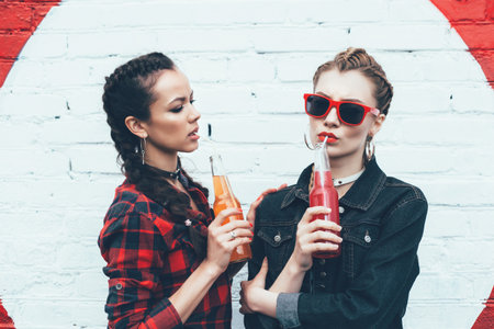 Two young women drinking colorful cocktails from bottles in the street. Fashionable style, hairstyle with braids. Outdoors lifestyle toned portraitの写真素材