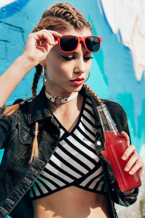 Young woman in sunglasses drinking colorful cocktails from bottles in the street. Fashionable style, hairstyle with braids. Close-up lifestyle portraitの写真素材