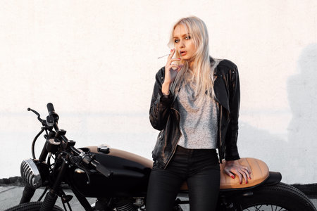 Outdoor lifestyle portrait of sexy biker girl wearing leather jacket sitting on a vintage custom motorcycle. Woman smoking cigaretteの写真素材