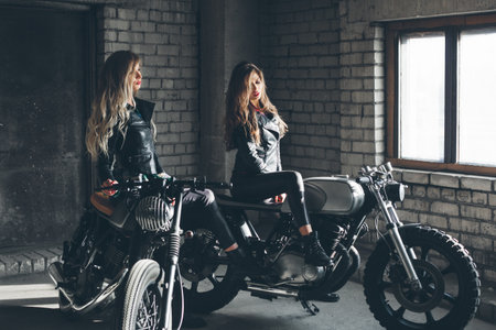 Sexy biker girls sitting on vintage custom motorcycles. Beautiful women with Healthy Long Hair ringlets wearing stylish leather jacketsの写真素材