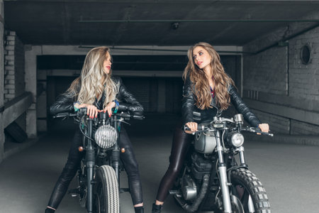 Sexy biker girls sitting on vintage custom motorcycles. Beautiful women with Healthy Long Hair ringlets wearing stylish leather jacketsの写真素材