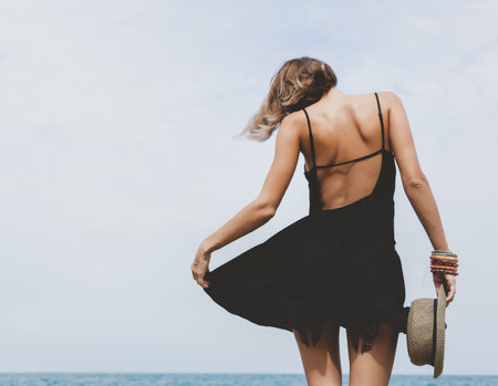 Outdoors lifestyle portrait of a girl dressed in a black dress with a hat in her hand standing on the background of the seaの写真素材