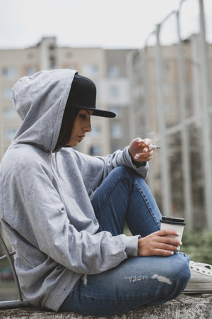 Outdoors lifestyle portrait os young woman sitting on the ground in the square, smoking a cigarette and drinking coffee from a paper cup. Hoodie, cap, jeans, snickers swag girl conceptの写真素材