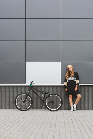 Outdoor lifestyle portrait of pretty sexy young girl in hockey jersey style dress posing on gray wall background with street bicycle. Copyspace availableの写真素材