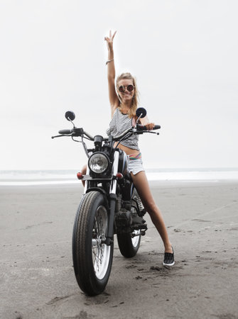 Girl in jeans shorts sitting on vintage custom motorcycle traveling and having fun, young pretty blonde woman posing near ocean, beach baliの写真素材