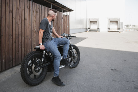 Handsome rider guy with beard and mustache sitting on classic style cafe racer motorcycle at day time. Bike custom made in vintage garage. Brutal urban lifestyleの写真素材