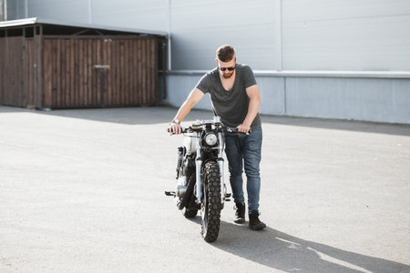Handsome rider guy with beard and mustache pushing classic style cafe racer motorcycle next to him at day time. Bike custom made in vintage garage. Brutal urban lifestyleの写真素材