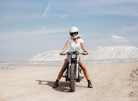 Sexy female rider in helmet sitting on her custom built cafe racer motorcycle in the desertの写真素材