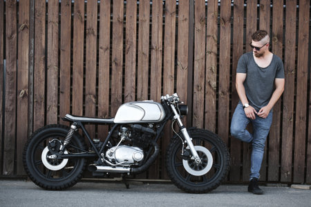 Handsome rider guy with beard and mustache standing next to classic style cafe racer motorcycle near wooden wall at day time . Bike custom made in vintage garage. Brutal fun urban lifestyleの写真素材