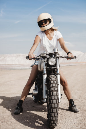 Sexy female rider in helmet sitting on her custom built cafe racer motorcycle in the desert