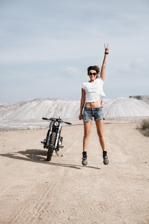 Young pretty cheerful woman jumping with raised hand. Girl biker with perfect fit slim tamed body and custom cafe racer motorbike on the desert. Outdoor lifestyle portraitの写真素材