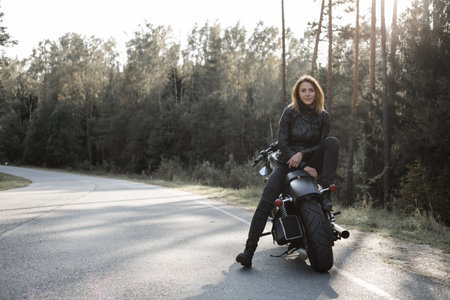 Young pretty cheerful woman sitting on motorcycle. Girl biker with perfect fit slim body and custom chopper motorbike at forest. Outdoor lifestyle portraitの写真素材