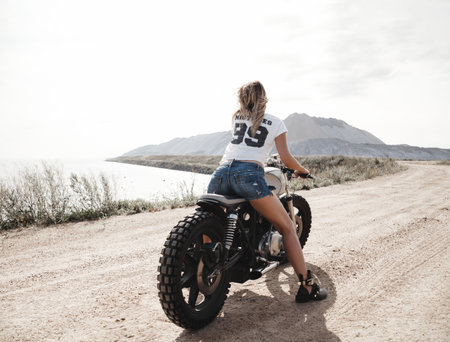 Young pretty cheerful woman sitting on custom cafe racer motorbike on the desert. Girl biker with perfect fit slim tamed body and long hairs. Outdoor lifestyle portraitの写真素材
