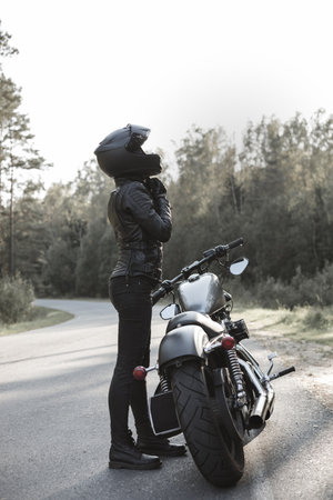 Young pretty cheerful woman putting on motorcycle helmet. Girl biker with perfect fit slim body and...