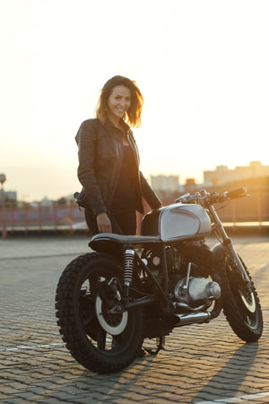 Biker woman ridding custom cafe racer motorcycle on empty city road at sunsetの写真素材