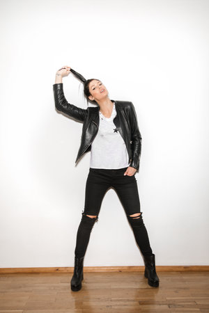 Attractive young girl in black ripped jeans and black leather jacket standing having lowered one hand in a pocket another hand raises her tail upの写真素材